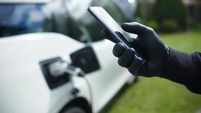 A black gloved hand holds a mobile phone near a white electric car which is charging