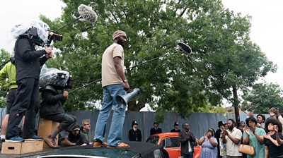 Behind the scenes with an actor being filmed standing on the roof iof a car