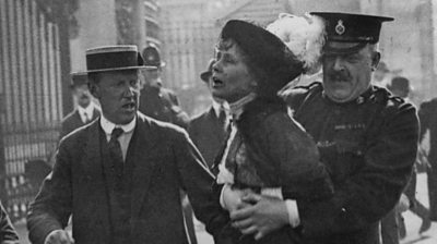 Policeman clasps Emmeline Pankhurst as another man walks along them