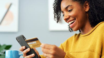 A person is holding a credit card and a mobile phone. She is smiling