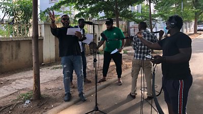 Five people are gathered outdoors in a courtyard around  a set of microphones. On the far left, a man in a black T shirt and jeans wearing dark glasses gestures in the air as he reads a script. On the far right, a sound man holds a recorder and adjusts the microphone.