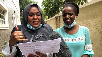 Two women are shown at a microphone, outdoors against a beige wall. One is wearing a grey headscarf and gesturing as she reads from a script. To the right, a second woman in a green dress grimaces as she looks on.