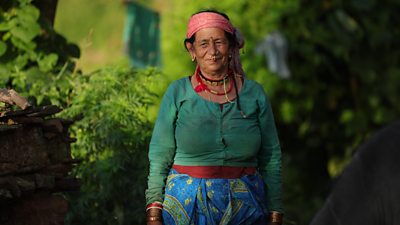 A woman in traditional Nepalese dress faces the camera. She is set against a backdrop of green vegetation.