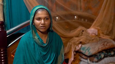 A young woman, Nazma Begum, wearing a blue is shown sitting in her home facing the camera in Dhaka, Bangladesh. A  bed, made with a mosquito net, is behind her in the small room.