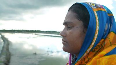 Fisherwoman Shabjan Begum, wearing a blue and gold headscarf and looking serious, is shown in profile against the sea near her home in Choto Antighara, Bangladesh.