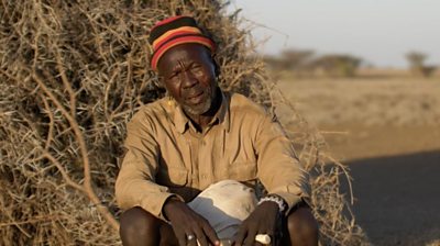 A man in tan shirt and brightly striped hat is shown leaning against a building made of thatched branches in northern Kenya.