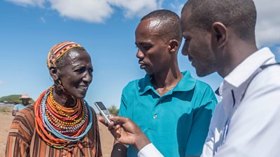 Two men interview a woman in traditional colourful clothing in rural northern Kenya. One is holding a small recorder. The landscape is dry.
