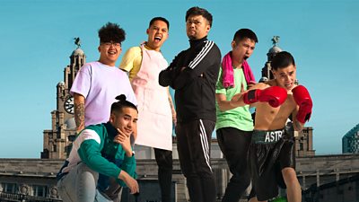 Six young males posing for the camera in front of the Royal Liver Building. One is wearing boxing attire, and one a pink apron.