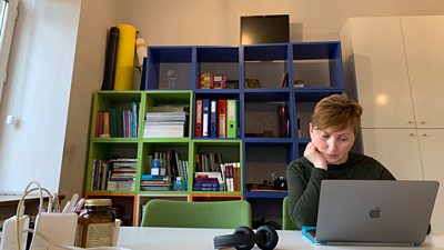 A woman sits at a table  in front of a Mac laptop with her head resting on her hand. Her headphones are off to the side on the table.  Behind her is a blue and green painted bookshelf, with books neatly arranged.