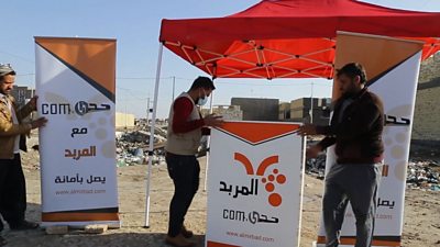 three men carrying signs saying 'al mirbad,com' in arabic set up a podium under an orange awning. they are in southern iraq with dry bare soil on the ground.