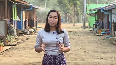 a young burmese woman with long dark hair wearing a grey shirt and purple patterned skirt faces the camera, behind her is a dusty road and rows of makeshift homes and shops.