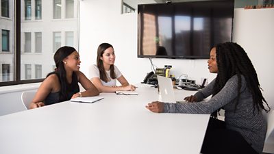 Three women - two black and one white - in a meeting in a generic white conference room with a window to the left and a blank TV screen on the wall.
