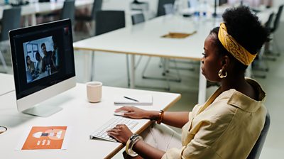 Black women in a yellow top with a tellow scarf around her hair at a computer in a generic hotdesk area.