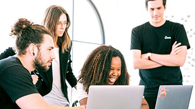 L-R: White man sitting with beard and a bun at a laptop, white women with long hair and glasses standing behind a black women with long hair smiling while looking at a laptop. Final hite man standing up with arm folded looking at laptop.