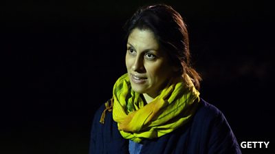 A woman in a blue jumper and yellow scarf around her neck looks up - set against a black background
