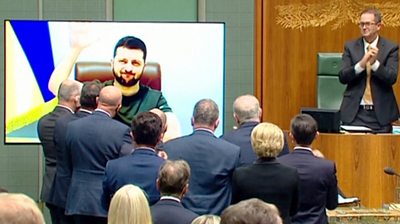 Australian parliament standing up and clapping at a TV screen with President Zelensky on