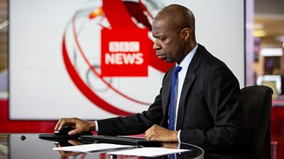 Side profile of Clive Myrie sitting in the 鶹ҳ News at Six studio. He's looking down, holding a mouse in his right-hand and wearing a suit with a blue tie, In the background is the 鶹ҳ News ident.