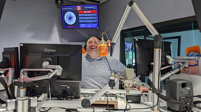 Presenter Wynne Evans sits in a Radio Wales studio in front of an orange mic, which he is laughing into