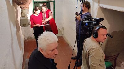 Two women dressed in red review a script on a clipboard while three men - one behind a camera with headphones on - look on. They are in a small concrete room under bright lights.