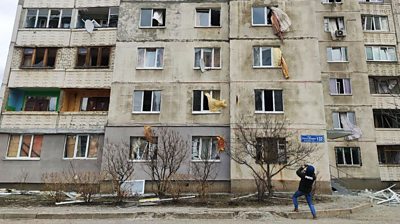 A cameraman films an apartment building with blown-out windows in Kharkiv, Ukraine. Glass and metal litters the ground around him.