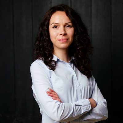 A woman with brown hair wearing a light-blue shirt with buttons stands against a dark background with arms folded.