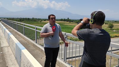 A man in a grey polo shirt and dark trousers holds a red microphone as he talks to a man filming him with a TV camera. They are on a bridge overlooking a large field with mountains in the distance.