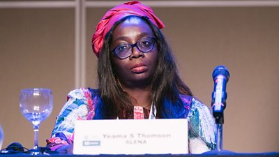 A Black woman wearing a blue and pink top and pink hair wrap is seated at a table looking at the camera. She has glasses, is behind a microphone and a card in front reads Yeama S Thompson, SLENA