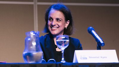 A woman with black hair smiles as she looks slightly away from the camera. She is wearing a black top and is seated at a table behind a microphone and pitcher and glass of water. The card in front of her reads Sheetal Vyas,