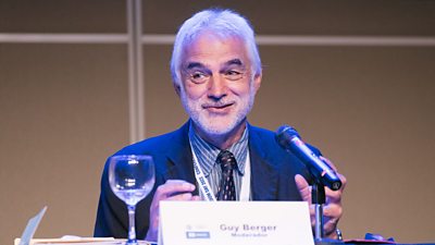 A white man with white hair and white beard gestures while sitting at a microphone behind a table. He wears a striped shirt, dark patterned tie and dark blue jacket.