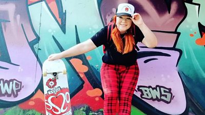 Woman poses with skateboard next to graffiti wall