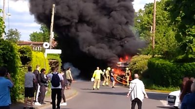 A bus engulfed in flames with thick black smoke as people watch firefighters