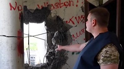 A wall with a hole blasted through it, a man is pointing at it