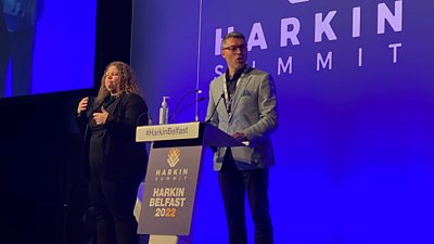 Adam Smyth at the podium on the Harkin Summit main stage. Blue background. He is wearing a grey blazer and blue trousers, glasses. To his left is a woman with long blonde hair doing BSL.