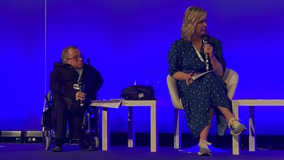 Allan MacKillop in a wheelchair holding a mic while on the main stage at Harkin Summit. To his left is Tara Mills sitting down in a green spotty dress. They're looking to their left and the background is a blue screen,