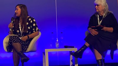 Sharmin Rahman left with a mic sitting in a white chair. On the right is Fiona Keane looking to her. There's a white table between them and a blue screen behind them.