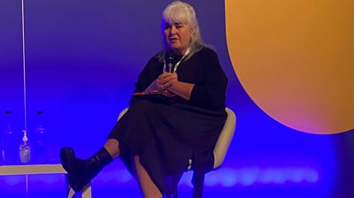 Fiona Keane wearing all back and white hair pinned back. Legs crossed in a white chair. Background blue with a orange circle on the right.