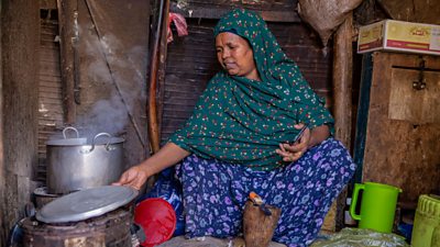 A woman in a green headscarf and blue dress sits in a simple wood-and-tin structure, taking a metal lid off a large pot on a low stove. A green jug is on the floor beside her.