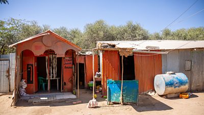 A small orange structure with a peaked roof, built of wood and tin and painted, sits on dry sandy ground, with another wood-and-tin structure adjacent and a dusty blue water tank in the front.