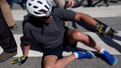 President Biden wearing a bike helmet. He is sitting on the ground, trying to get up.