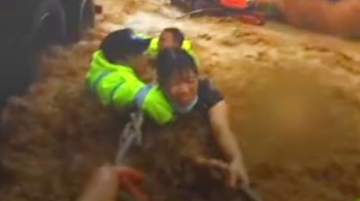 A woman holds onto a rope in rushing flood waters, with rescue workers holding her