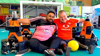 Omar Mehtab, wearing a pink football shirt and Paul Carter, wearing a red football shirt, sit between two orange robots.