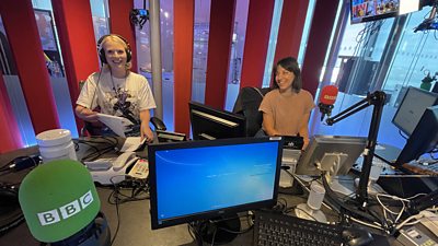 Two women sit in an audio recording studio at  Broadcasting house, there is a green microphone and a red microphone in the shot, with a computer screen and mixing desk. Maha Taki is recording on the red mic and wears a beige t-short, she has brown hair. Saskia Black is producing wearing headphones and a white t-shirt with a pattern in black, she is holding the script papers.