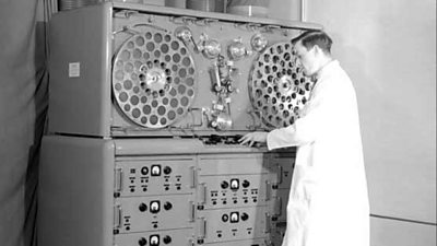 An engineer in a lab coat with 'VERA', a huge machine with two large tape drums