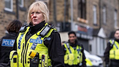 Sarah Lancashire as Catherine Cawood. She has a florescent police stab vest on, and has a bloodied face