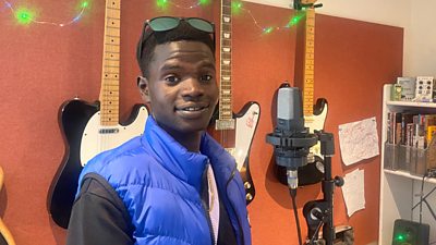 Mustafa stands in front of a microphone in a recording studio, looking at the camera with sunglasses on his head
