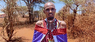 A young man in traditional colourful clothing faces the camera. He stands in front of a background of dry sand and trees without leaves.