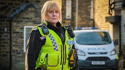 Sarah Lancashire as Catherine Cawood in Happy Valley. She stands in the road in a high vis police vest and uniform