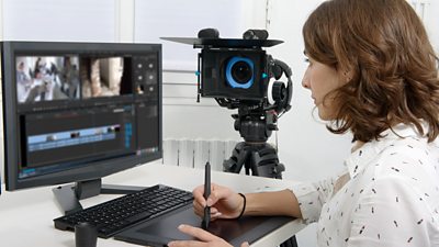 A woman edits video clips at her computer using a pen. A camera is in the background