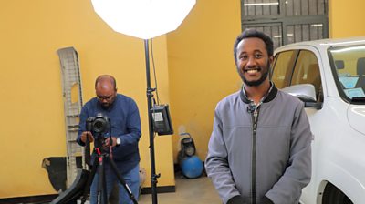 One man in gray jacket faces the camera, smiling, while another man sets up a videocamera.