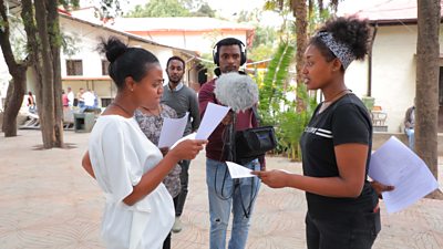 A young woman in white holds a script; a young woman in a black T shirt faces her also holds a script and is mid speech. Behind them a man holds a mic.